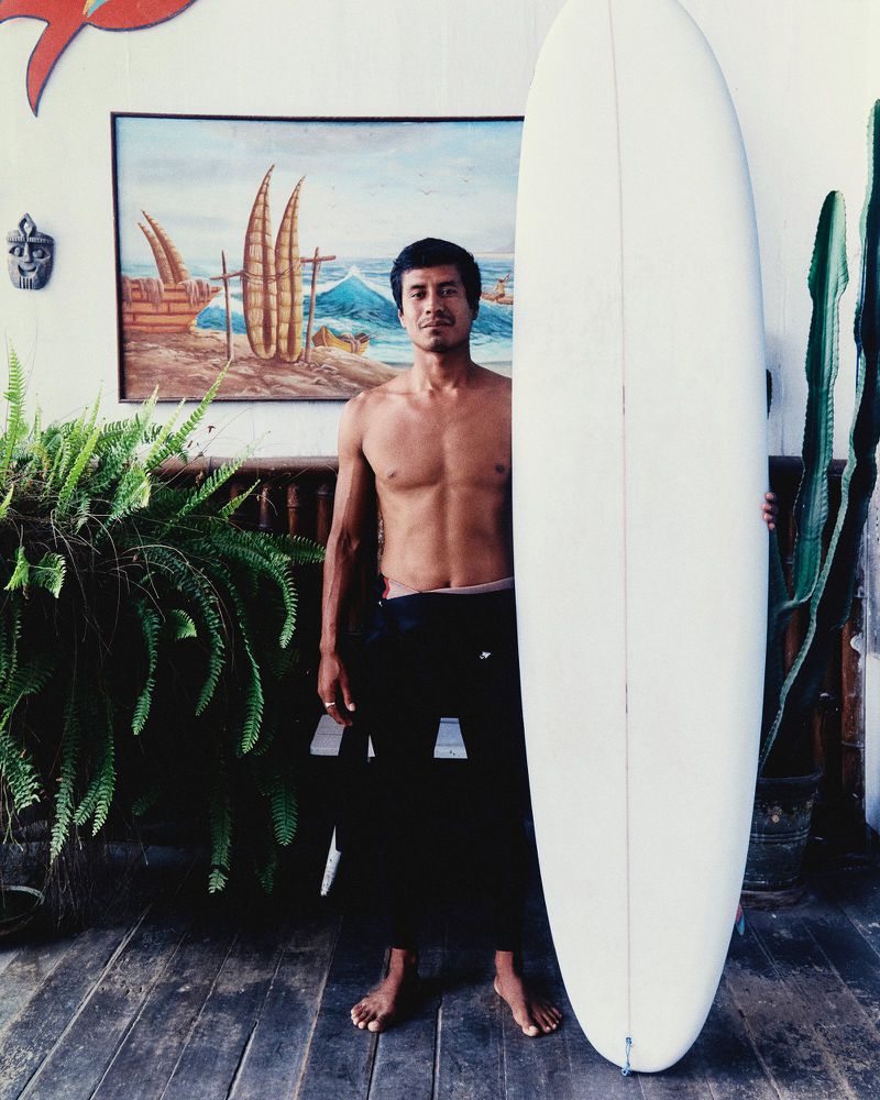 Shirtless man standing barefoot on a wooden deck holding a large white surfboard upright next to potted plants and a wall painting.