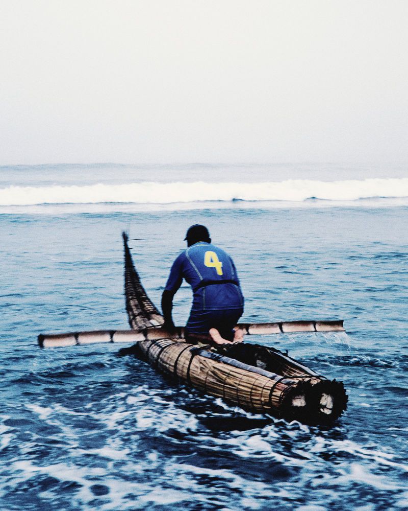 A person kneeling on a traditional reed boat holding a paddle while navigating through breaking ocean waves.