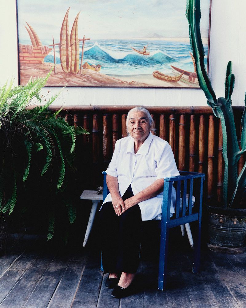 Elderly woman in a white short-sleeve shirt seated in a blue chair in front of a coastal painting and bamboo wall.