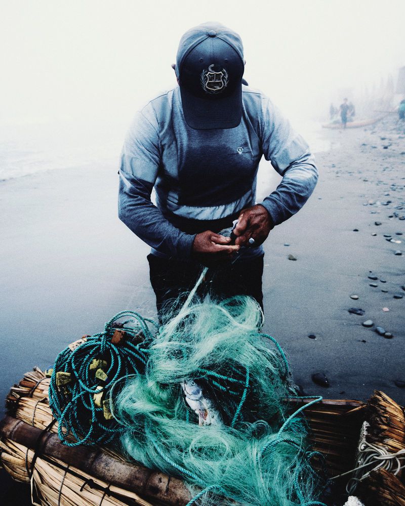Fisherman in a baseball cap untangling a green fishing net filled with catch over a traditional reed boat on a misty beach.