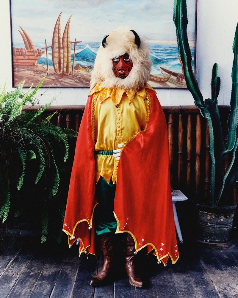 Figure in traditional Andean folklore costume with a horned fur mask, red scalloped cape, and yellow satin shirt on wooden flooring.