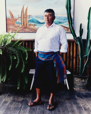 A man standing in a white button-down shirt, dark trousers, sandals, and a blue woven sash against a coastal painting backdrop.