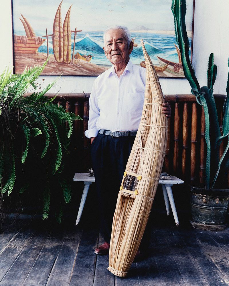 An elderly man in a white collared shirt stands holding a traditional woven totora reed boat model in front of a painted seascape.