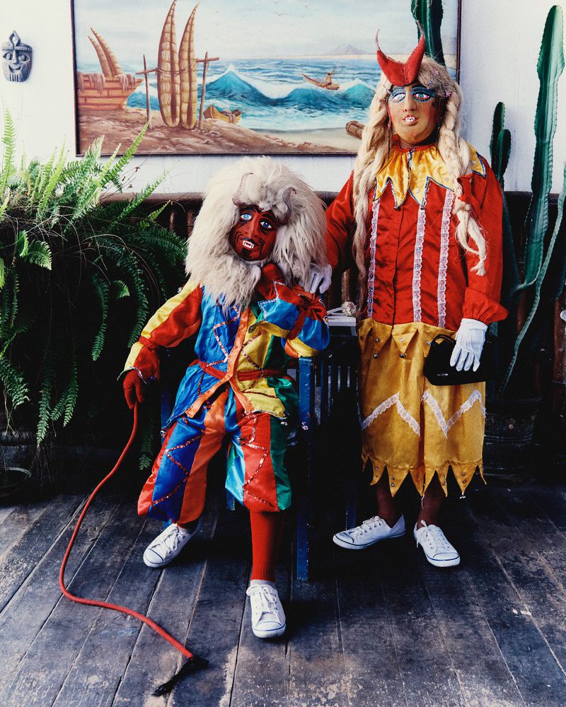 Two figures in colorful folkloric masks, wigs, and satin costumes posing on a wooden floor in front of a coastal painting.