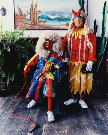 Two figures in colorful folkloric masks, wigs, and satin costumes posing on a wooden floor in front of a coastal painting.