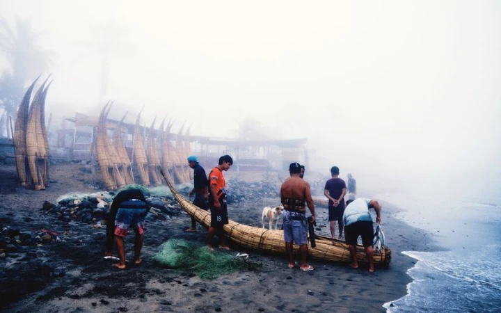 Fishermen gathered around traditional reed boats on a misty beach with fishing nets on dark sand.