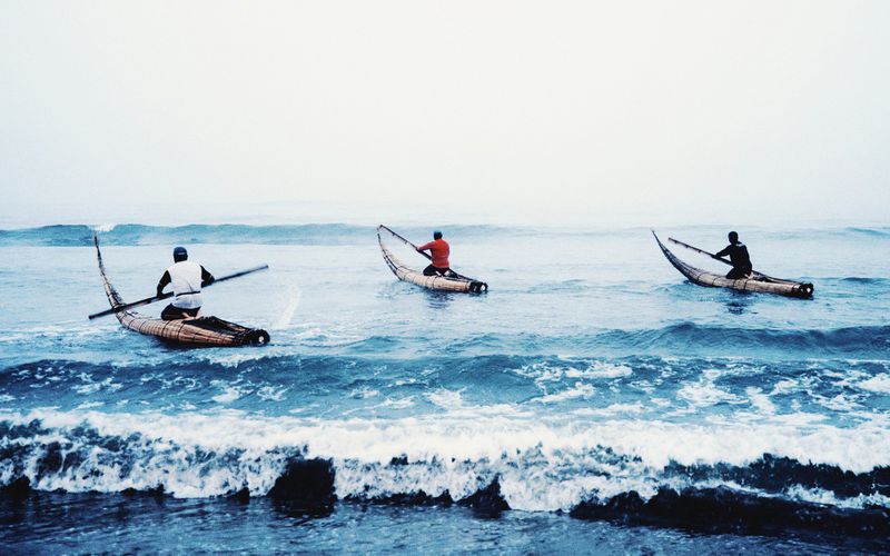 Three fishermen paddling traditional reed watercraft through ocean surf under an overcast sky.