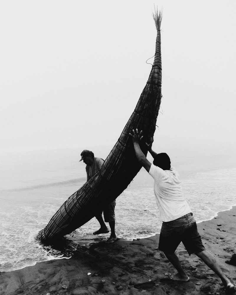 Black and white photo of two men lifting a large traditional woven reed boat on a sandy ocean beach shore.