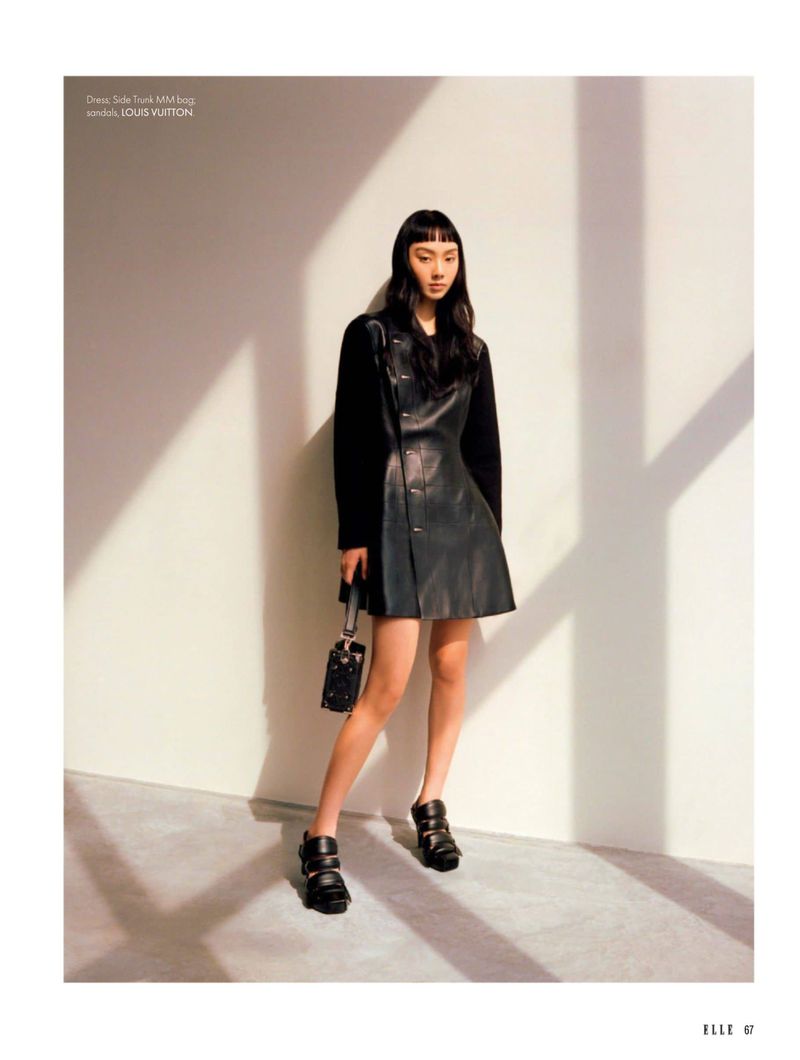 A fashionable young Asian woman poses indoors wearing a black leather dress, black boots, and carrying a patterned handbag in a fashion editorial shot.