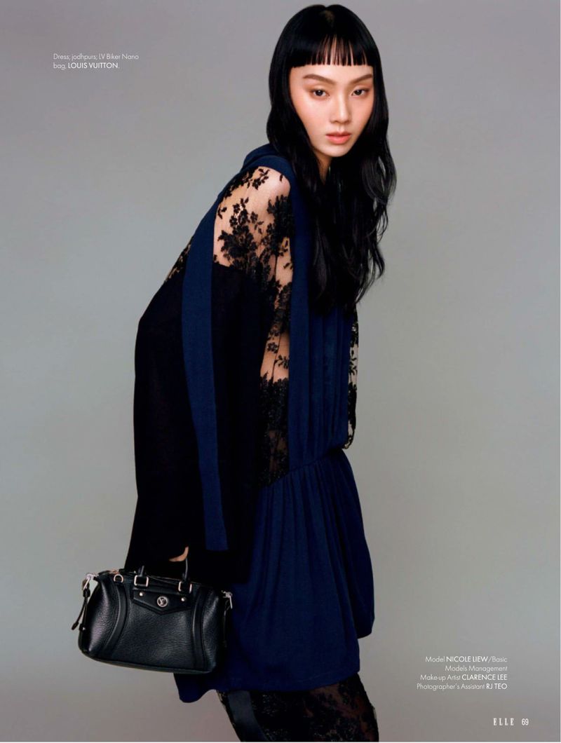 An Asian fashion model poses indoors in a dark blue dress with lace sleeves while holding a black leather handbag, against a grey backdrop.