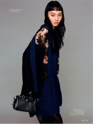 Model Nicole Liew in a dark blue dress with sheer lace sleeves holding a black handbag against a grey studio backdrop