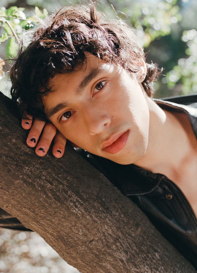 Young man with dark curly hair rests on a tree trunk in natural light. He wears a black jacket and has nail art on his fingers.