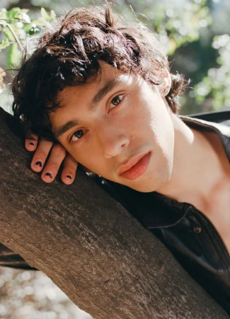 Young man with dark curly hair rests on a tree trunk in natural light. He wears a black jacket and has nail art on his fingers.