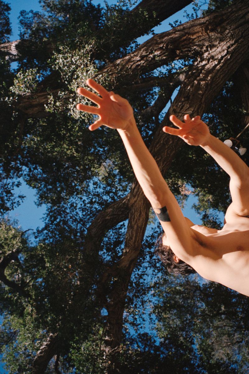 Low-angle shot of a shirtless adult man with arms raised toward the sky and tree branches in an outdoor setting on a sunny day.