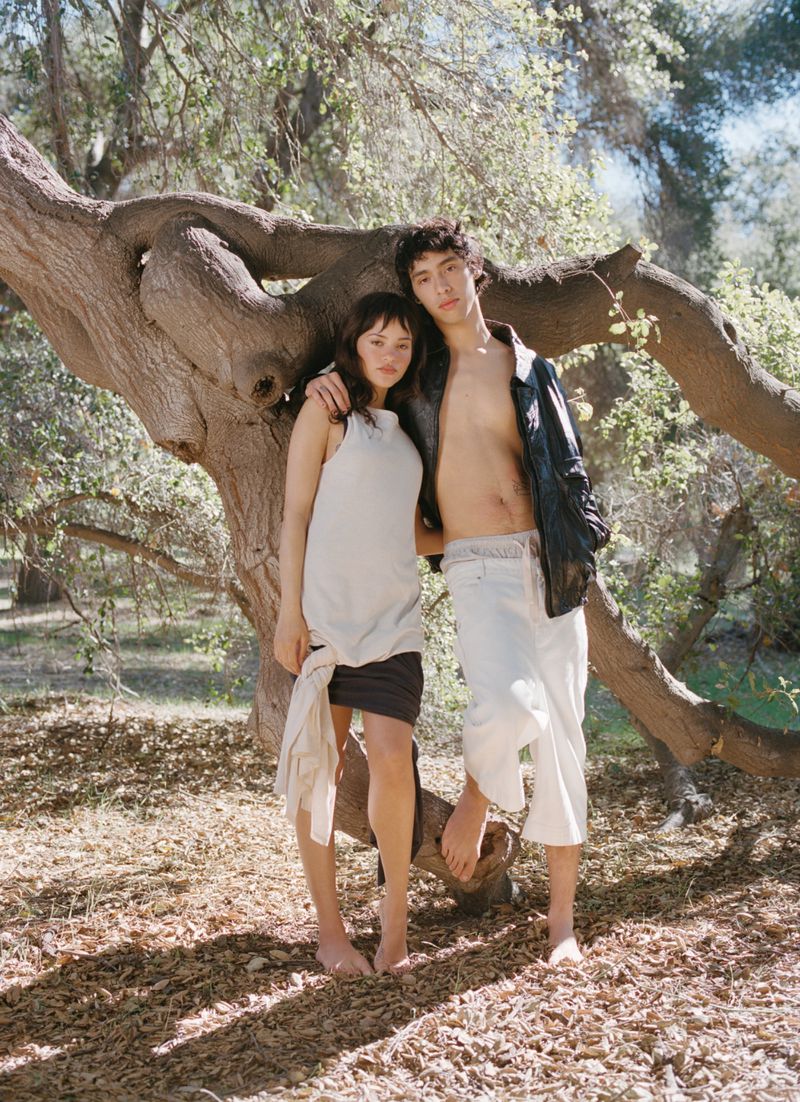 A young couple stands embracing next to a large tree in a sun-dappled forest, wearing minimal casual clothing, posing intimately.