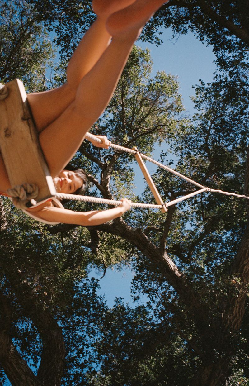 A woman swings playfully from a rope swing, captured from a low angle with trees and blue sky framing the scene.
