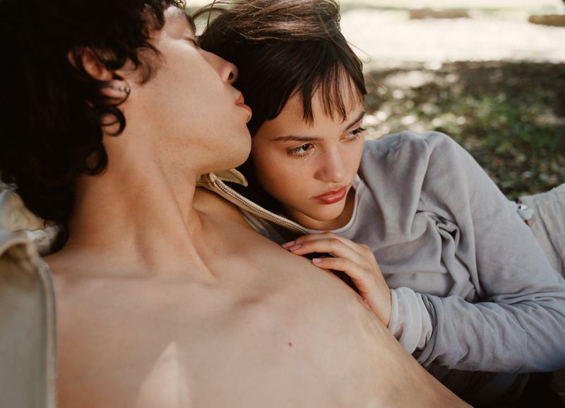 Intimate portrait of a young couple lounging outdoors; woman with hand on man's chest, both in natural light with a soft, romantic aesthetic.