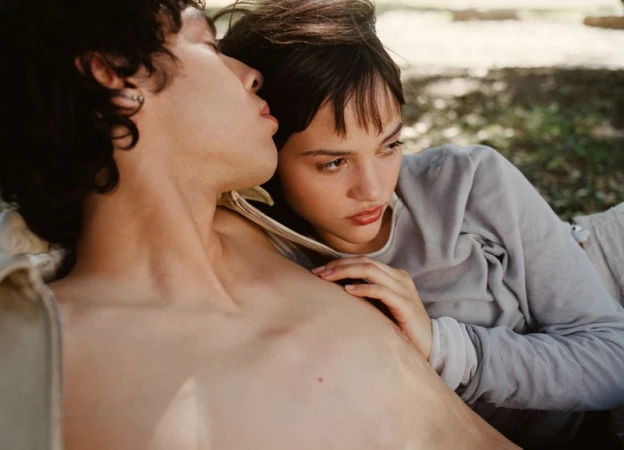 Intimate portrait of a young couple lounging outdoors; woman with hand on man's chest, both in natural light with a soft, romantic aesthetic.