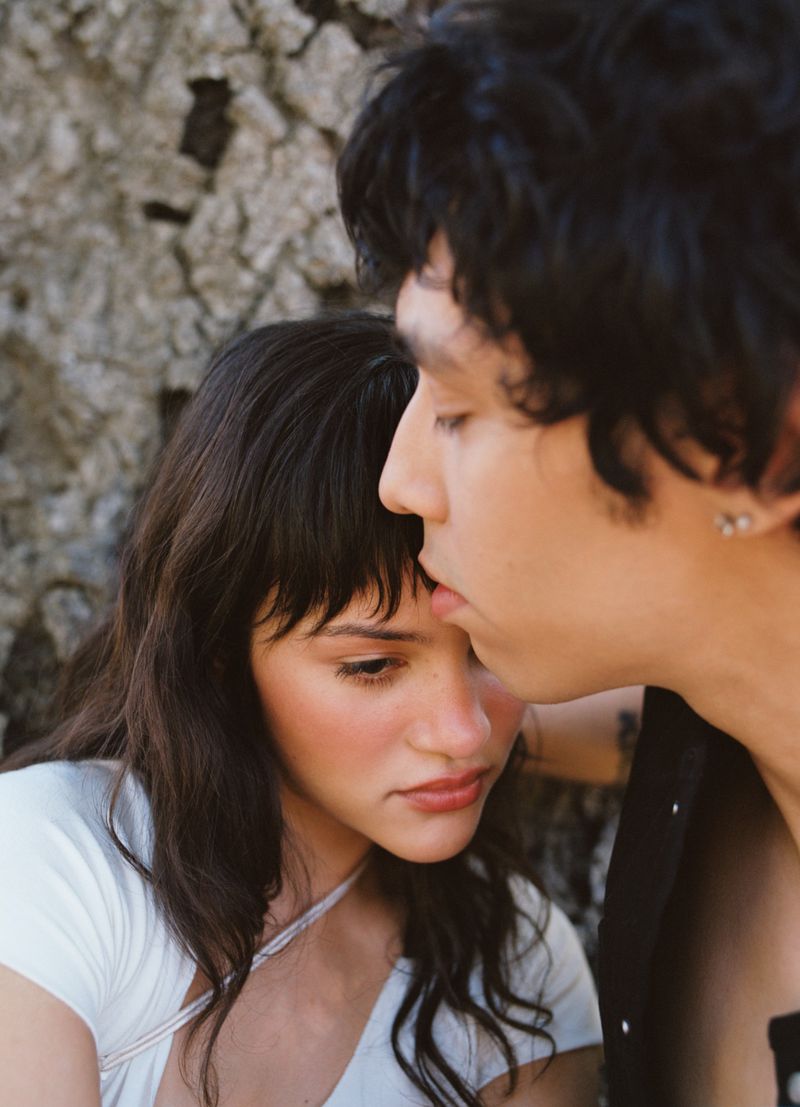Close-up of an adult couple embracing near a textured wall, bathed in soft natural light. The man kisses the woman's forehead.