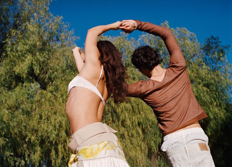 Two people dance together outdoors with trees and a blue sky in the background. One person wears a white top and floral skirt.