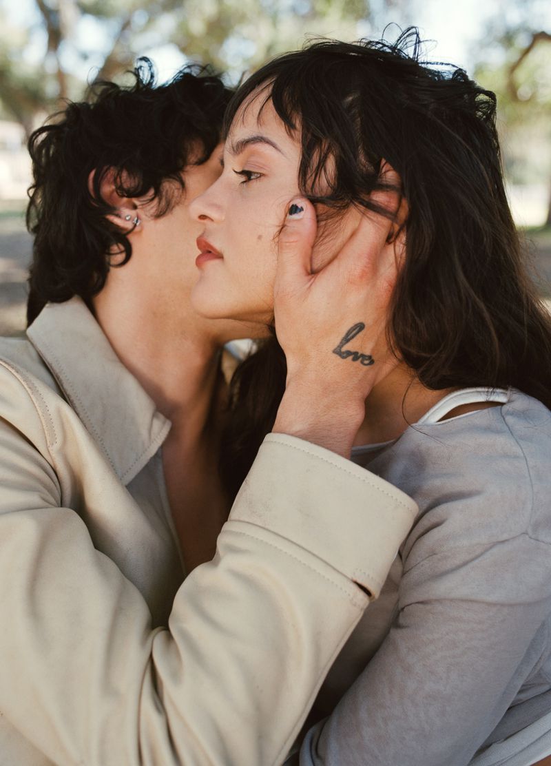 Close-up of a young, light-skinned heterosexual couple embracing in an outdoor setting. The woman has a tattoo on her forearm.