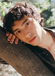 Close-up portrait of a young man resting his head against a thick tree trunk in a natural outdoor setting.