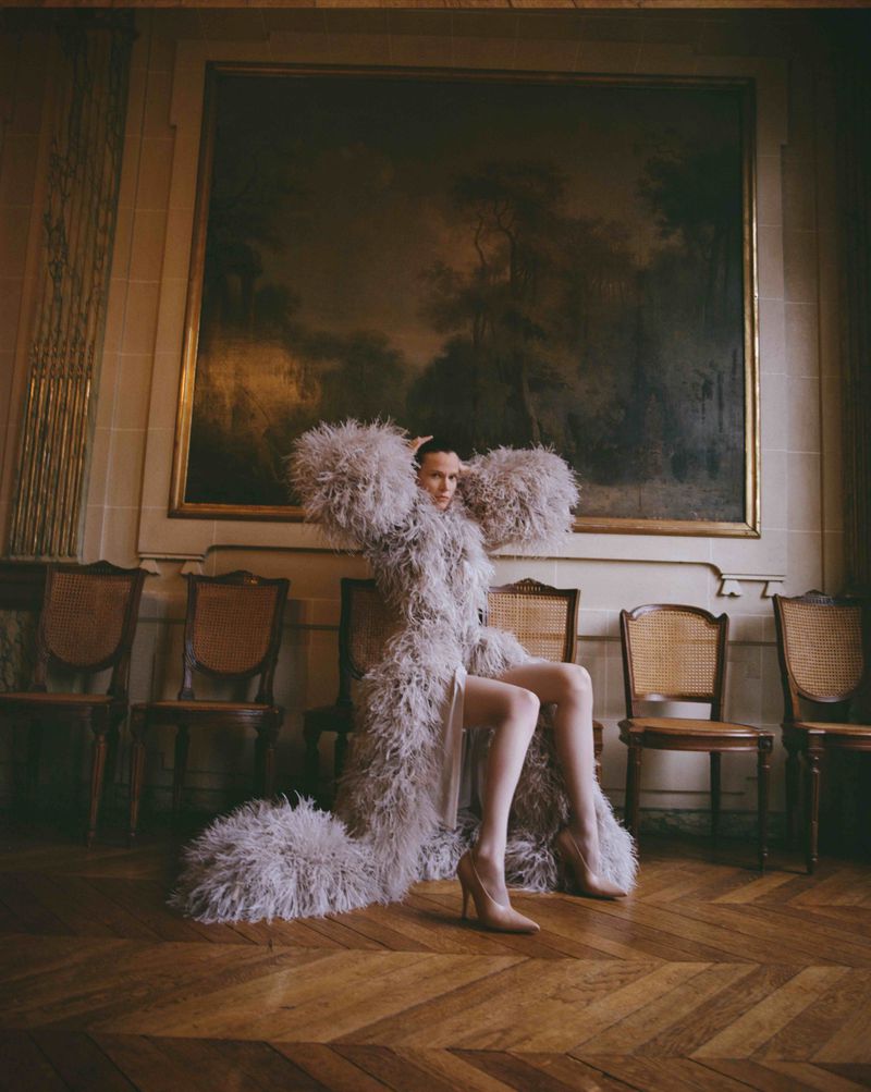 Model Santa Valentina seated in a voluminous feathered grey coat against a classical framed painting on chevron parquet flooring.