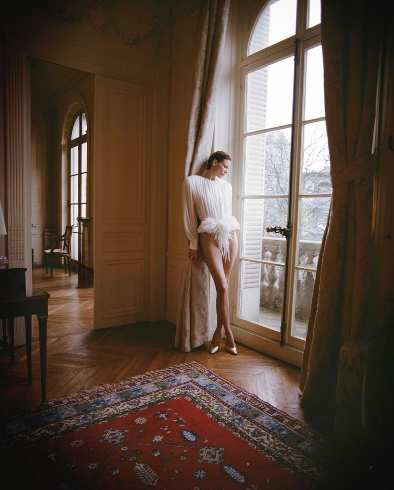 Model Santa Valentina posing by a tall arched window wearing a pleated white bodysuit in an ornate room with parquet floors.