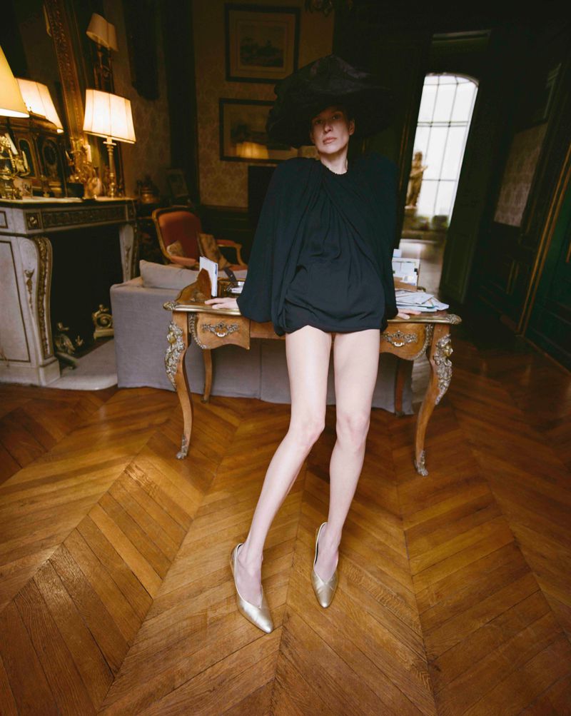 Model Santa Valentina in a voluminous black top, oversized wide-brim hat, and gold heels posing by an antique desk in an ornate room.