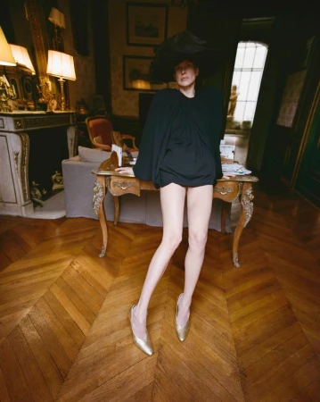 Model Santa Valentina in a voluminous black top, oversized wide-brim hat, and gold heels posing by an antique desk in an ornate room.