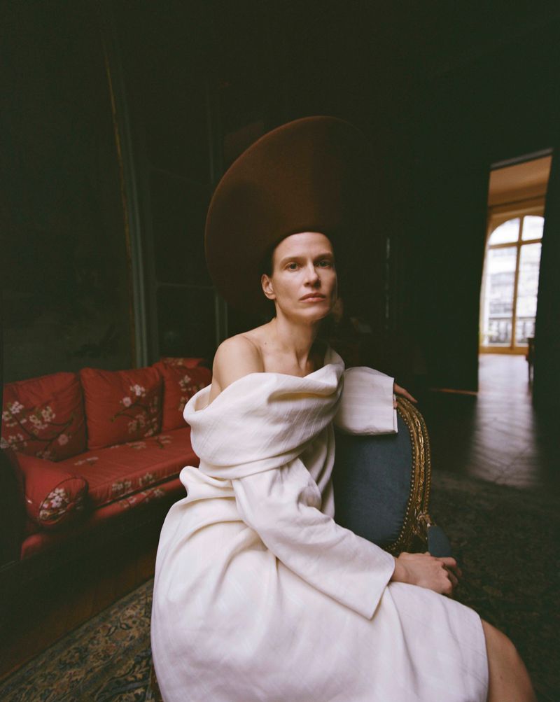 Model in an oversized brown saucer hat and off-the-shoulder white coat seated in an interior room on a vintage armchair.