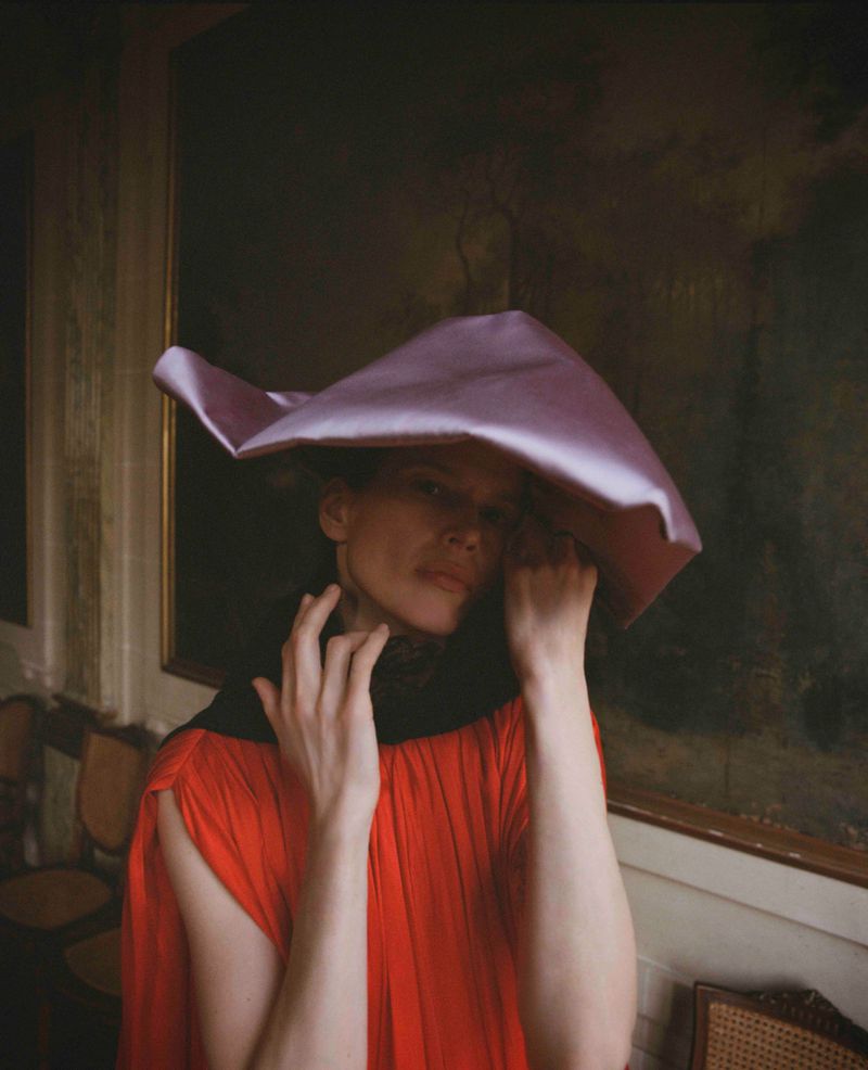 Model Santa Valentina in a sculptural lilac satin headpiece and pleated red dress against a dark framed painting.