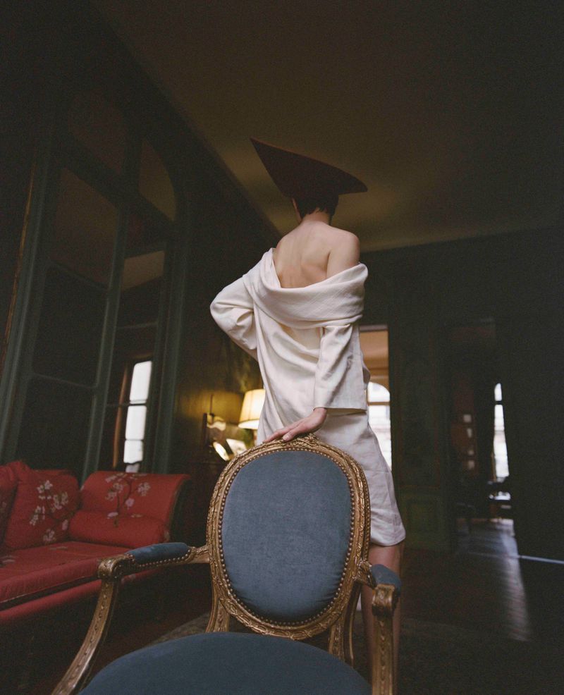 Model Santa Valentina standing from behind in an off-shoulder cream garment with a pointed headpiece beside an ornate armchair.