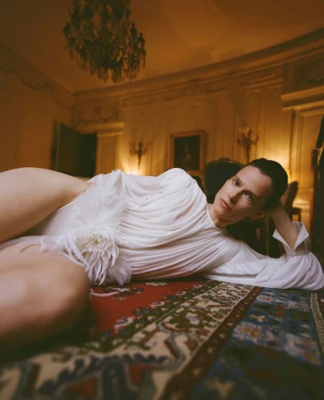 Santa Valentina reclining on an ornate patterned rug wearing a draped white dress in a warmly lit classic room.