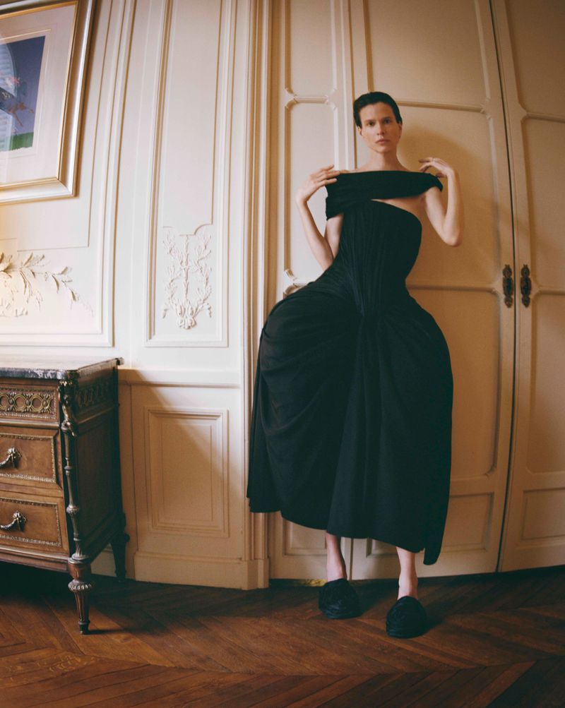 Model Santa Valentina wearing an exaggerated sculptural black dress standing in a room with parquet flooring and panelled doors.