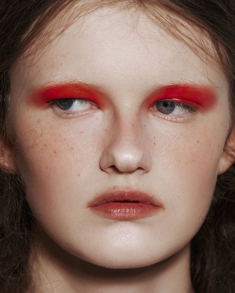 Close-up beauty portrait of model Aimee Byrne featuring bold, diffused red eyeshadow and faint eyebrows looking upward.