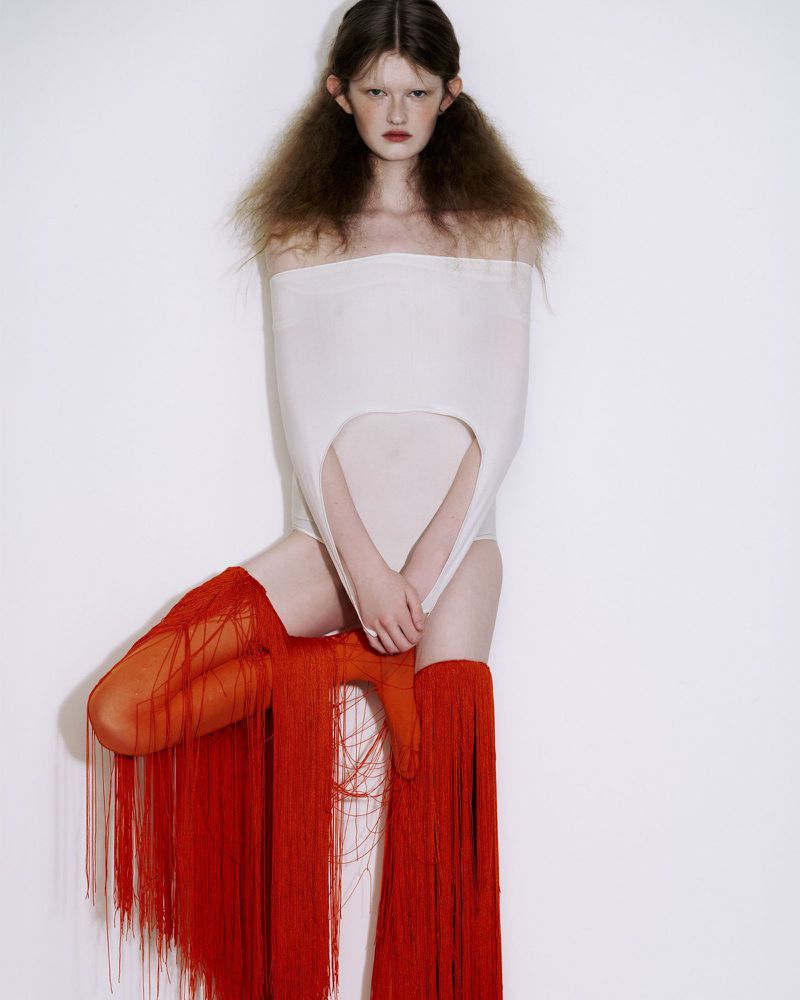 Woman in a white strapless bodysuit and red fringed hosiery poses against a plain white backdrop in a studio fashion editorial.