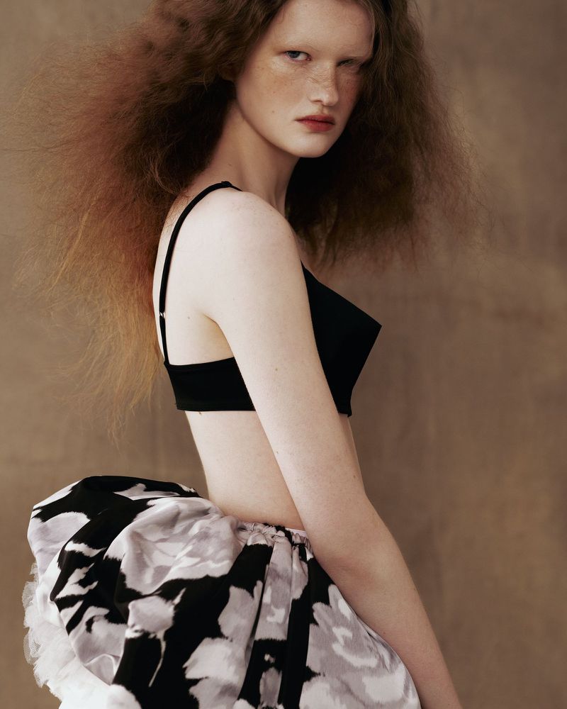 Model Aimee Byrne posing in a black crop top and monochrome floral puff skirt against a warm textured backdrop.
