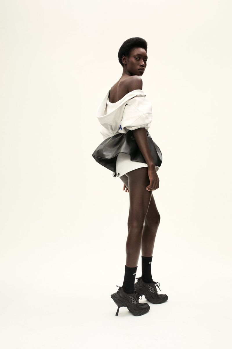 Fashion model in an off-the-shoulder top, skirt, shorts, socks, and sculptural heeled shoes posing in a brightly lit photo studio.