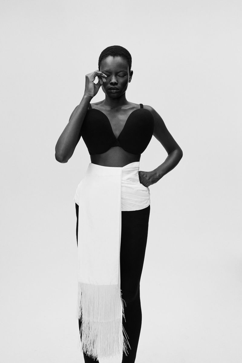 Portrait of a Black woman with short hair wearing a black bra, white fringed sash, and black leggings against a white backdrop.