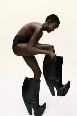 Dark-skinned woman in underwear puts on oversized black high heel boots in a fashion shot with a bright studio background.
