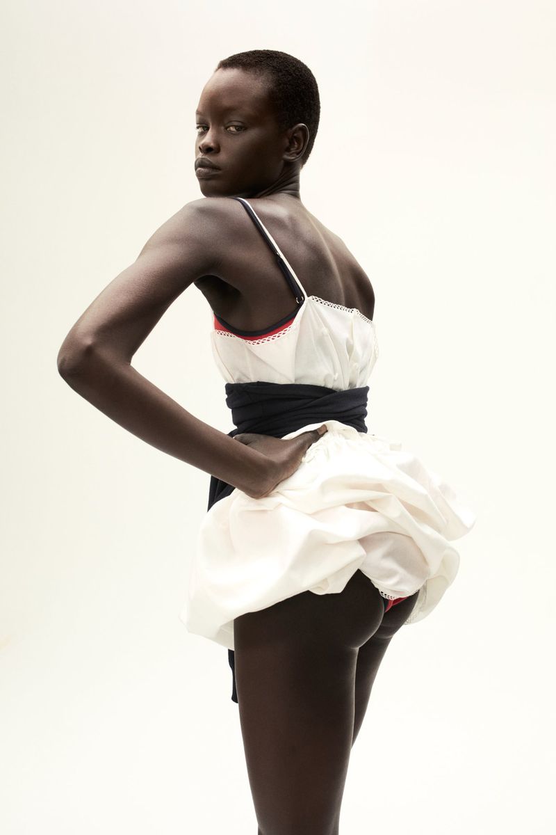 A young Black woman poses in a studio with a white dress and a black waist sash, looking over her shoulder in a fashion setting.