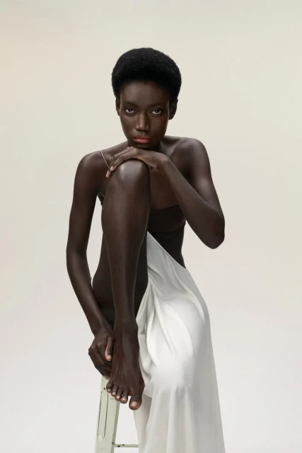 Portrait of a slender African American woman in a minimalist studio setting, draped in white fabric, seated on a distressed stool.