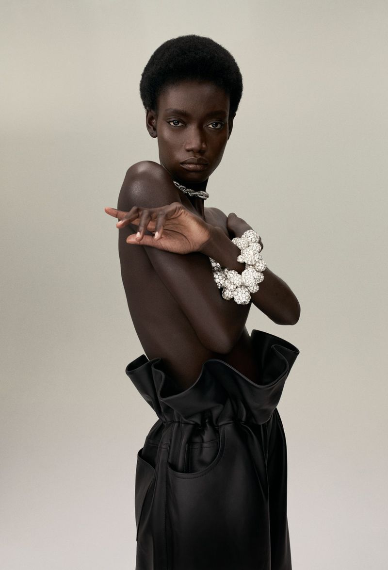 Dark-skinned woman poses in fashion shot, wearing black leather pants and shimmering, pearl-like jewellery against a pale backdrop.