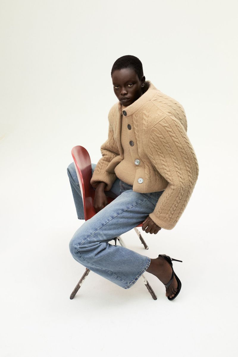 An adult female model sits on a red chair in a studio wearing a beige cardigan, blue jeans, and black heels, looking at the camera.