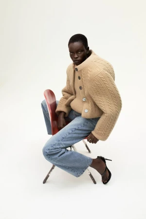 An adult female model sits on a red chair in a studio wearing a beige cardigan, blue jeans, and black heels, looking at the camera.