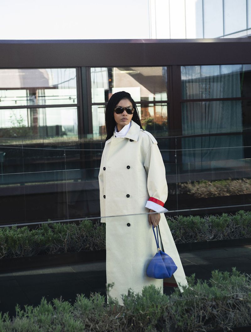 Sharon Alexie wearing a long off-white trench coat, sunglasses, and carrying a blue handbag outdoors.
