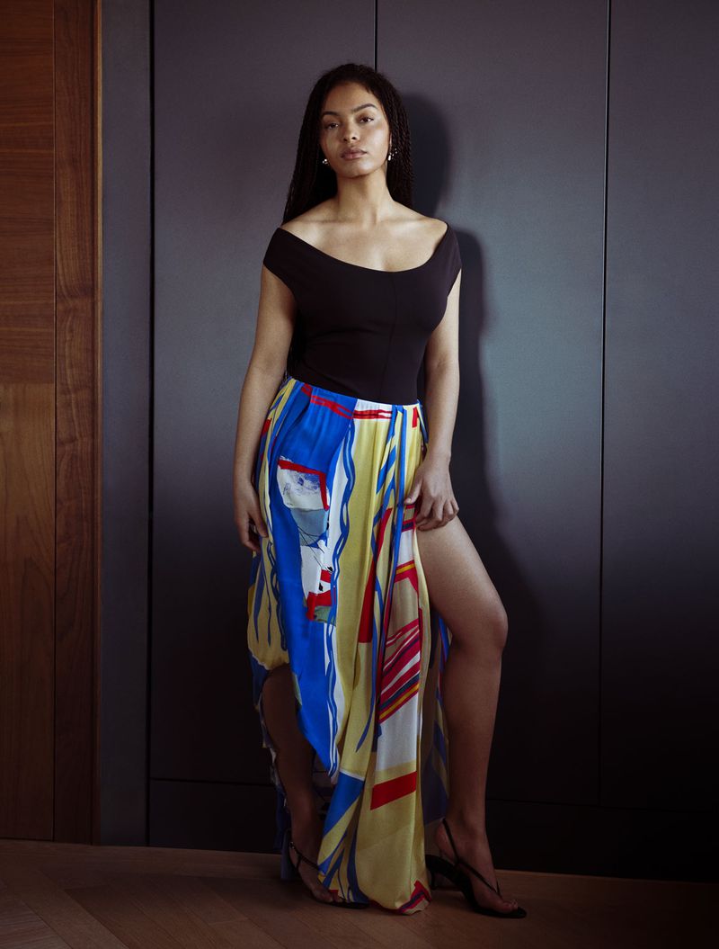 Model Sharon Alexie posing in a black off-the-shoulder top and colorful high-slit patterned skirt against dark wood wall paneling.