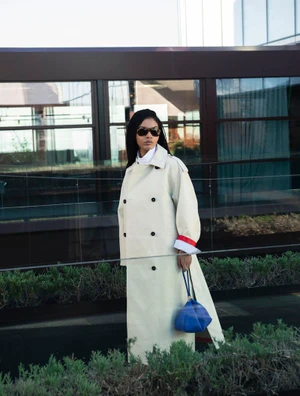 Sharon Alexie wearing a long off-white trench coat, sunglasses, and carrying a blue handbag outdoors.