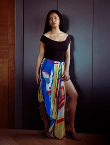 Model Sharon Alexie posing in a black off-the-shoulder top and colorful high-slit patterned skirt against dark wood wall paneling.
